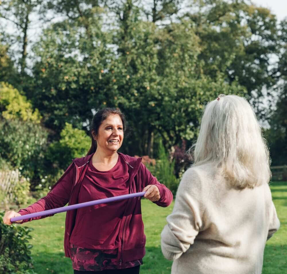 Two women outdoors, one holding an exercise band and smiling at the other. Trees and greenery are visible in the background. - Home Instead