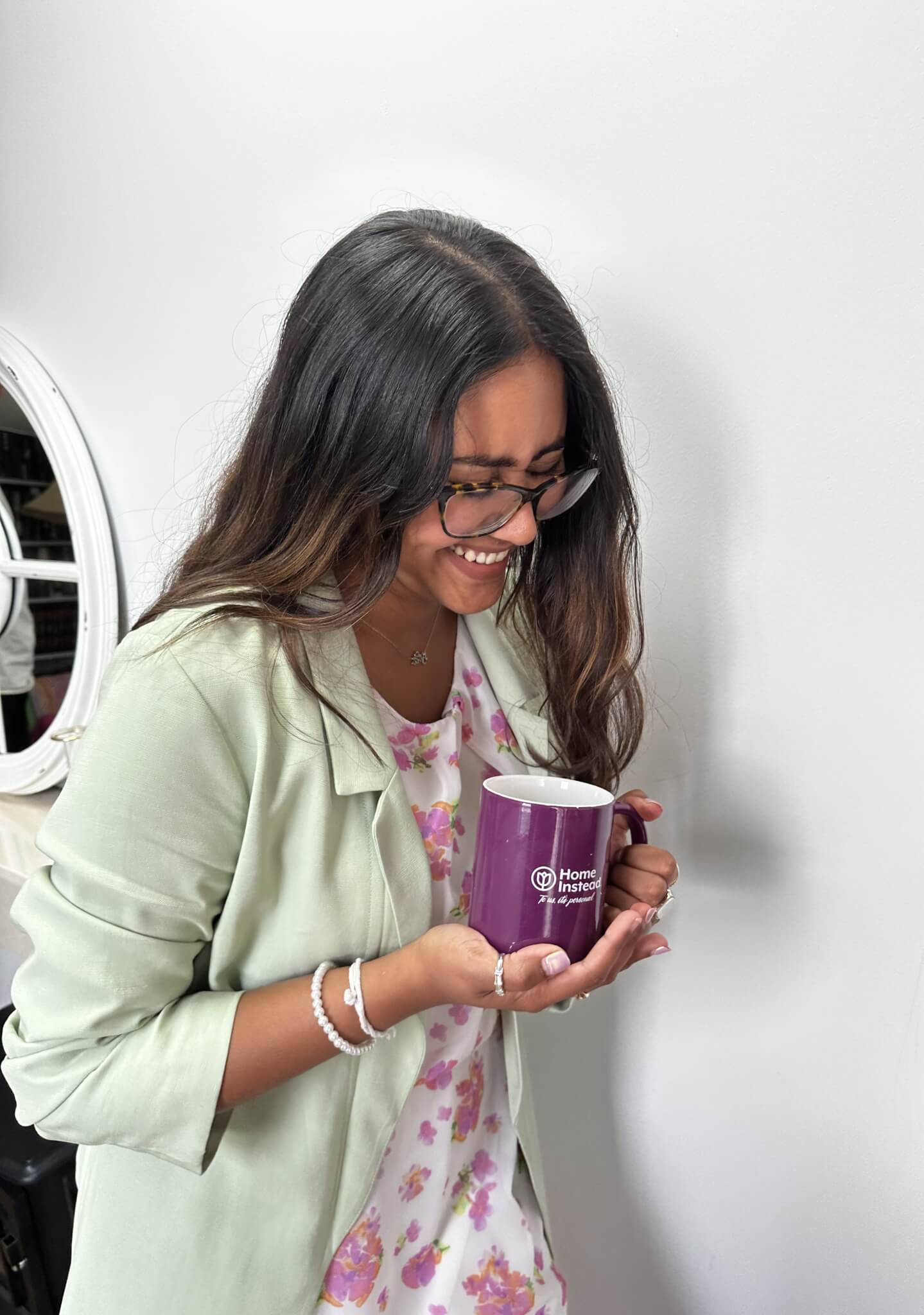 A woman with glasses, smiling and holding a purple mug, wearing a light green blazer over a floral dress. - Home Instead