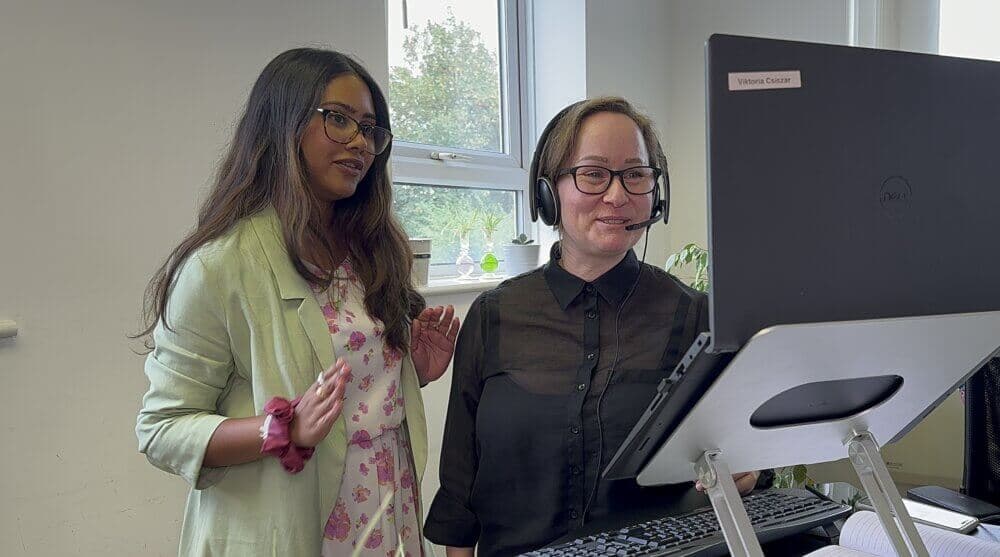 Two women smiling and conversing while looking at a computer screen, one standing and the other wearing a headset. - Home Instead