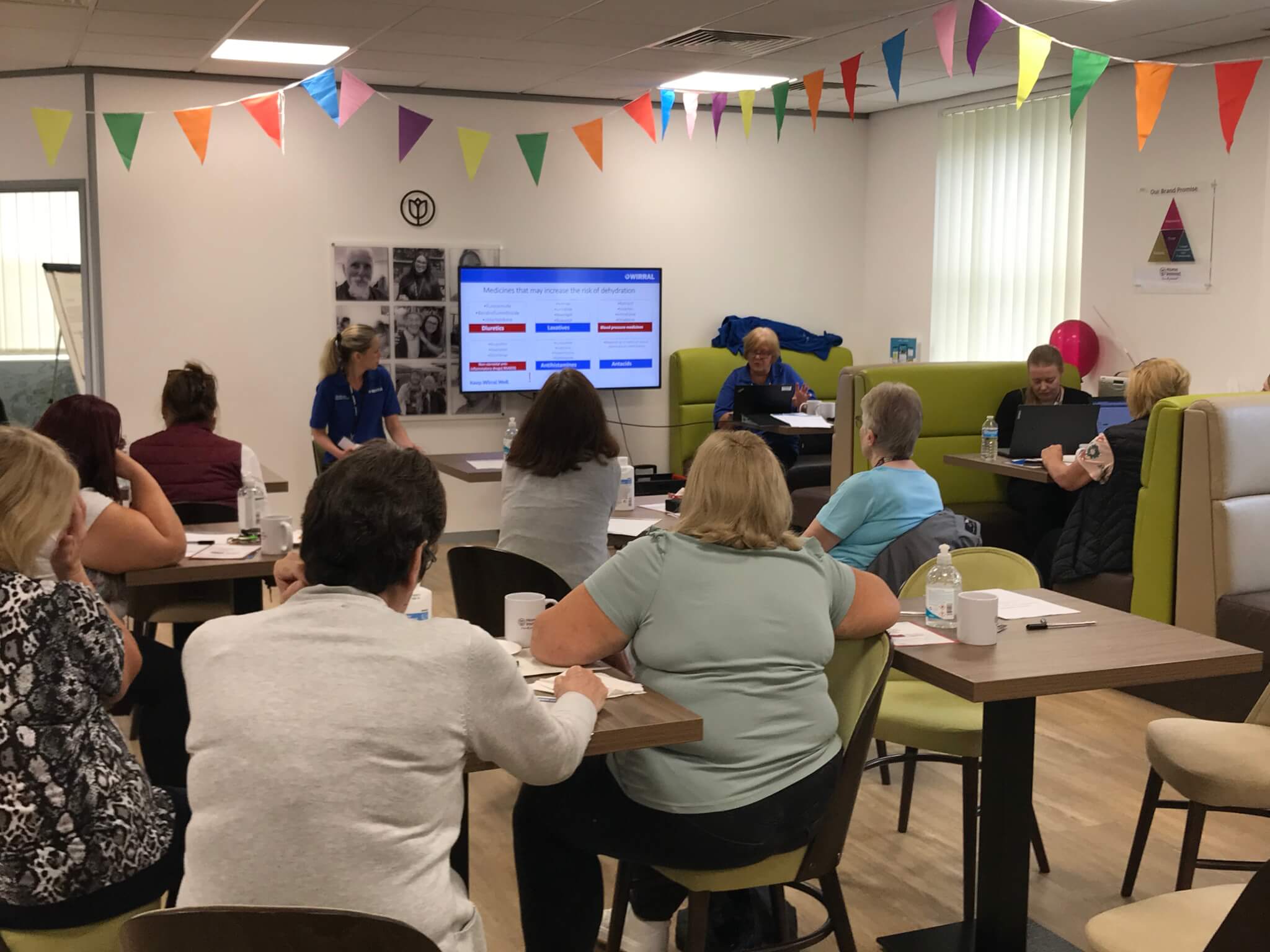 A group of people in a brightly decorated room attending a presentation with a screen displaying slides. - Home Instead