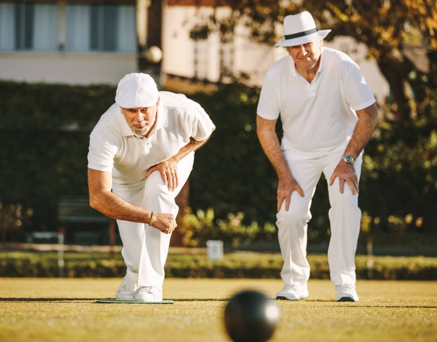 Two men in white attire playing lawn bowls; one rolling the ball while the other watches closely. - Home Instead