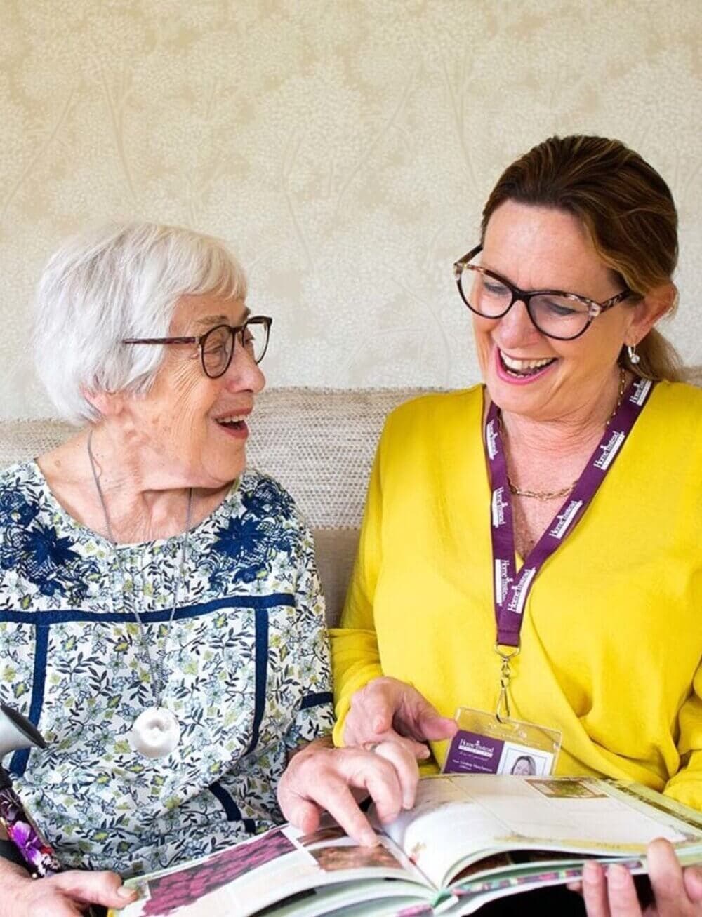 An elderly woman and a younger woman with a lanyard share a laugh while looking at a book together. - Home Instead Bournemouth & Christchurch