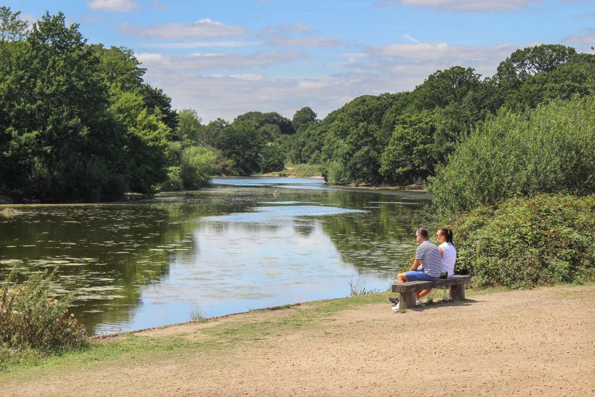 Two people sit on a bench near a calm river surrounded by lush greenery under a blue sky with scattered clouds. - Home Instead