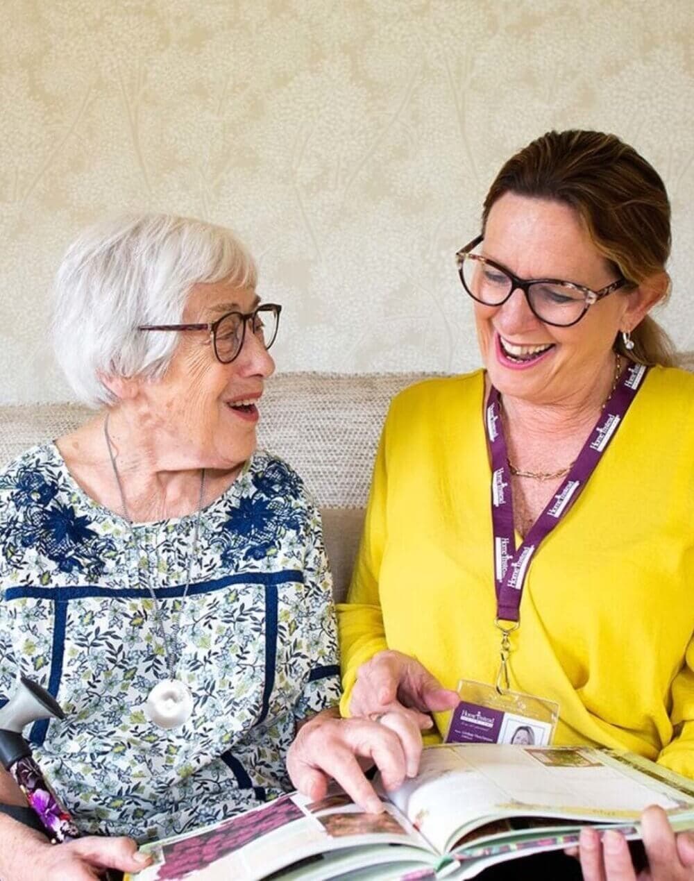 An elderly woman and a younger woman with a name badge smile while looking at a book together. - Home Instead