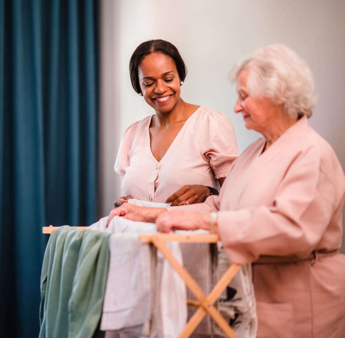A younger woman and an older woman smile while folding laundry together in a bright room. - Home Instead