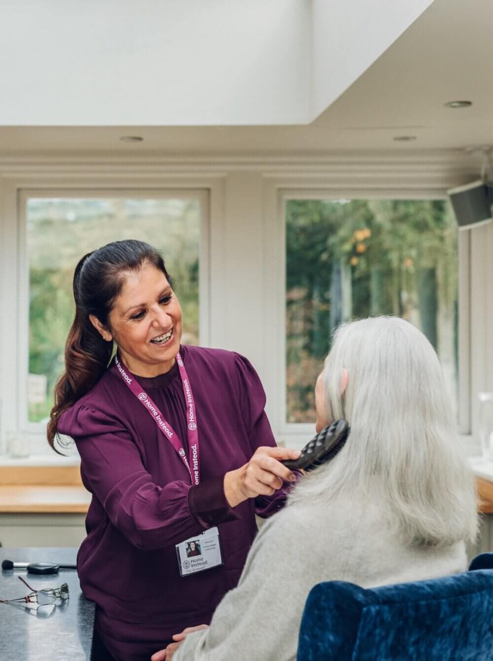 A caregiver in a purple top brushes the hair of an elderly person with long white hair in a bright room. - Home Instead