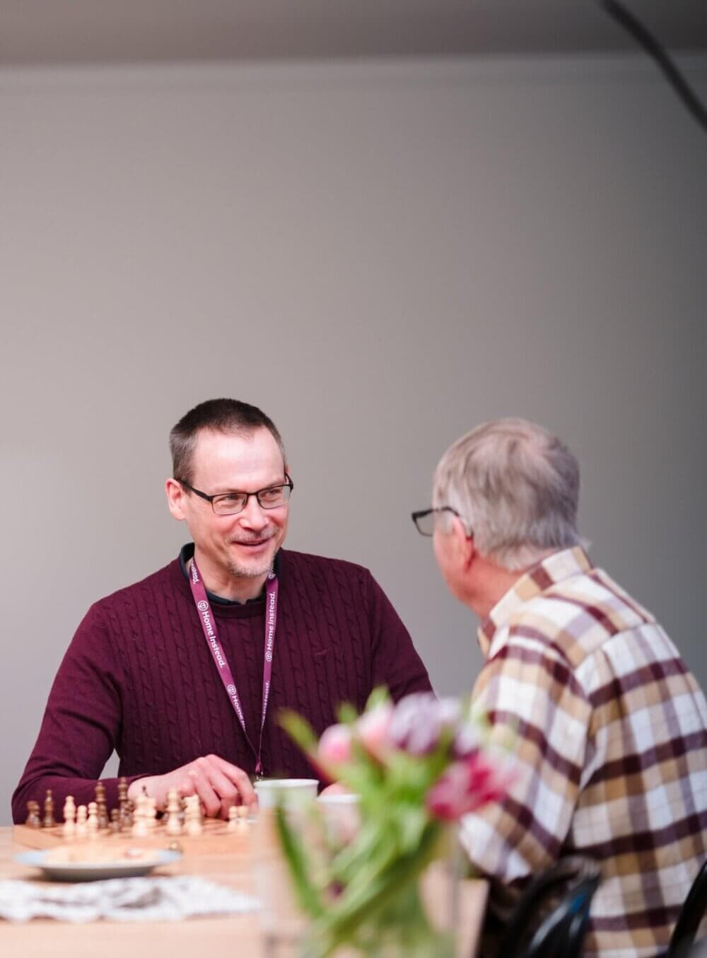 Two men sitting across from each other, engaged in conversation, with a chessboard and flowers on the table. - Home Instead