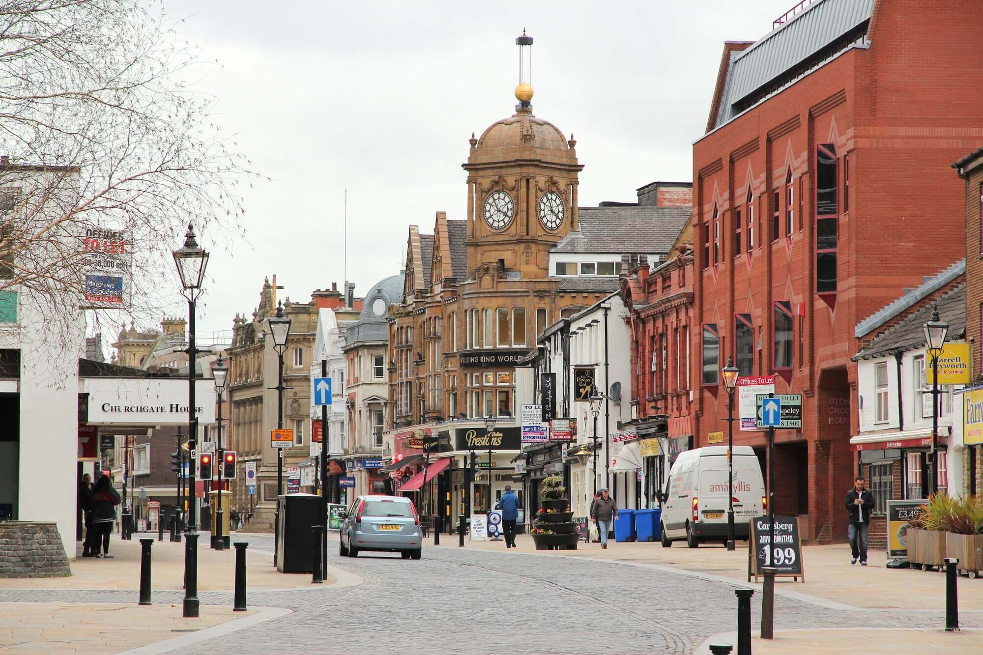 A bustling street scene with historic buildings, shops, a clock tower, cars, and people walking in an urban area. - Home Instead