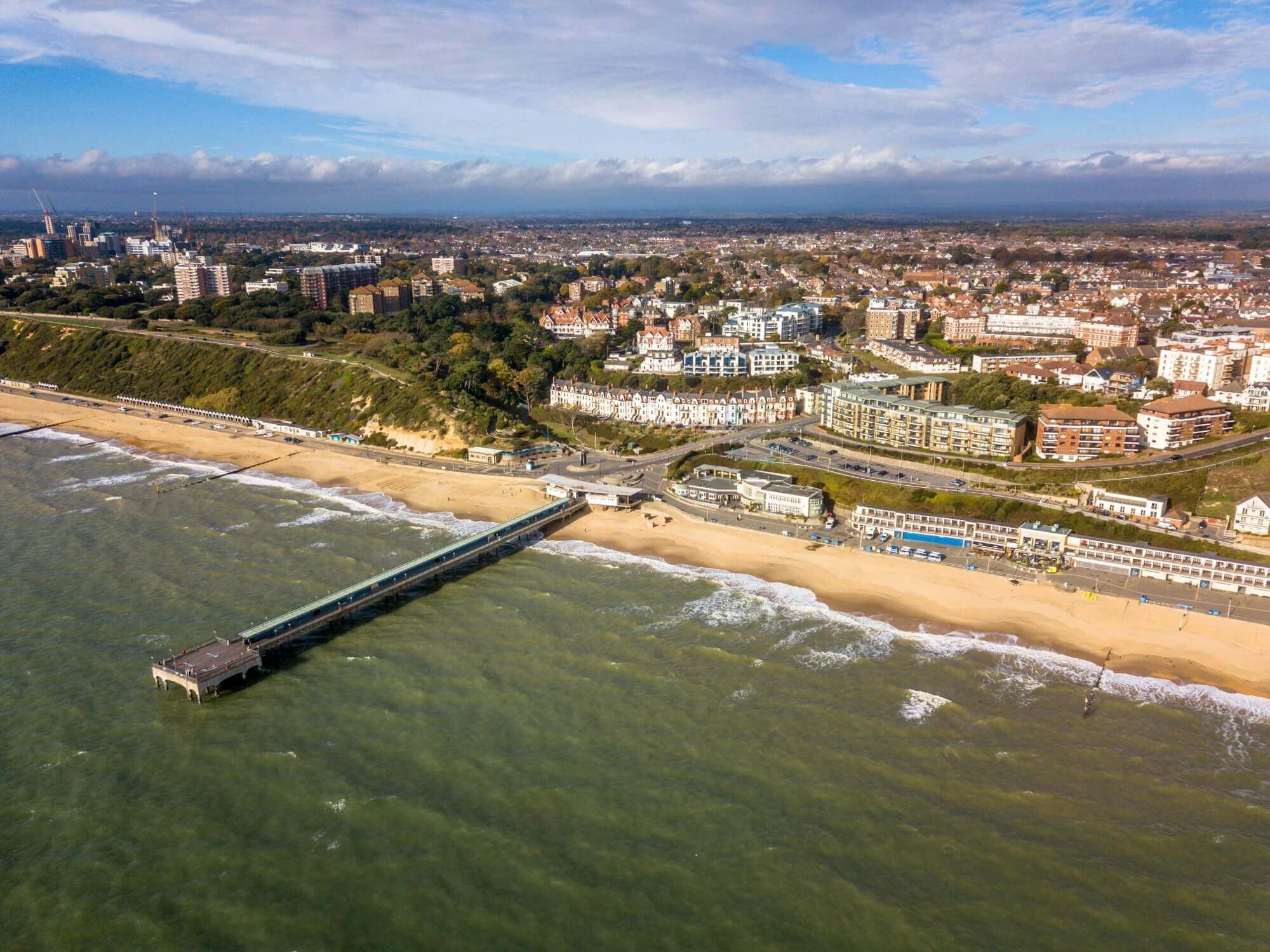 Aerial view of a coastal town with a long pier extending into the sea, sandy beach, and numerous buildings in the background. - Home Instead Bournemouth & Christchurch