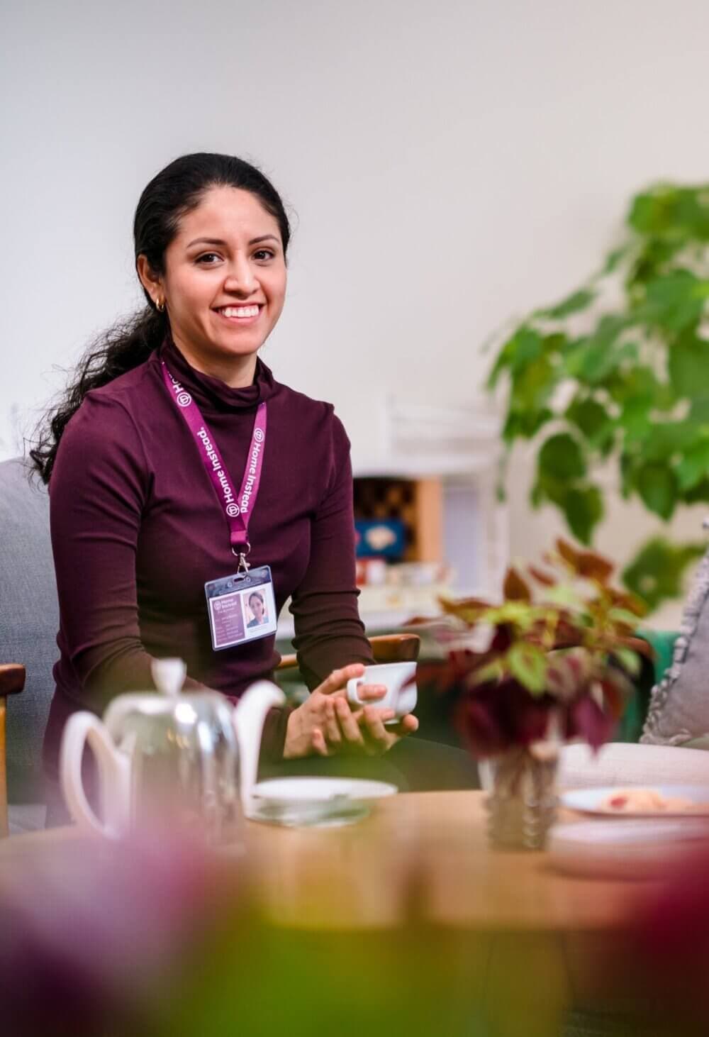 A smiling person wearing a purple shirt and ID badge holds a teacup while sitting at a table with a teapot and plants. - Home Instead