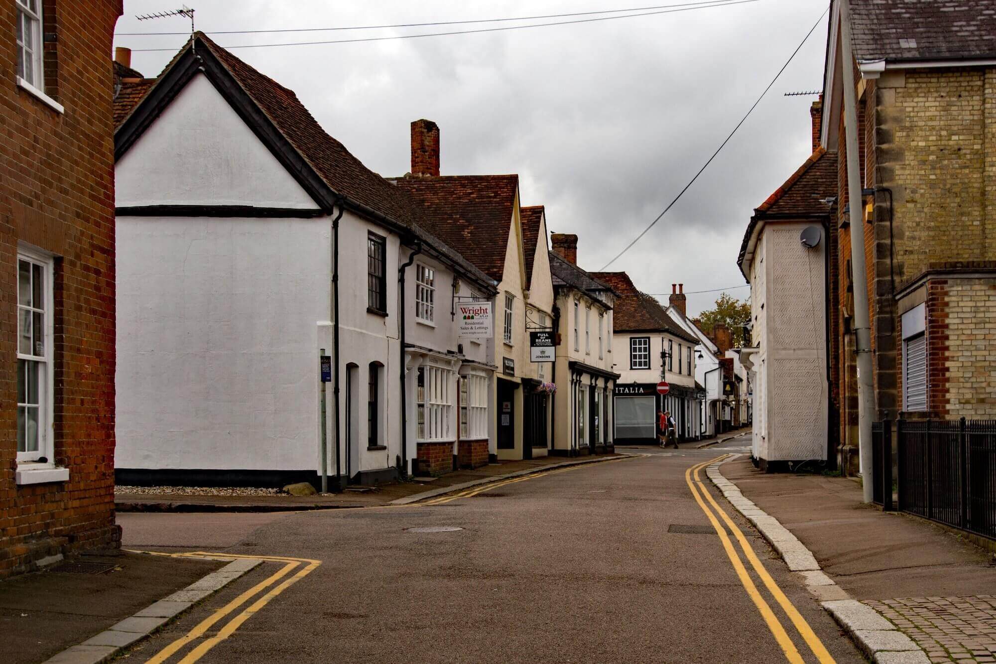 A narrow, empty street with old, white-painted buildings under a cloudy sky in a small town. - Home Instead