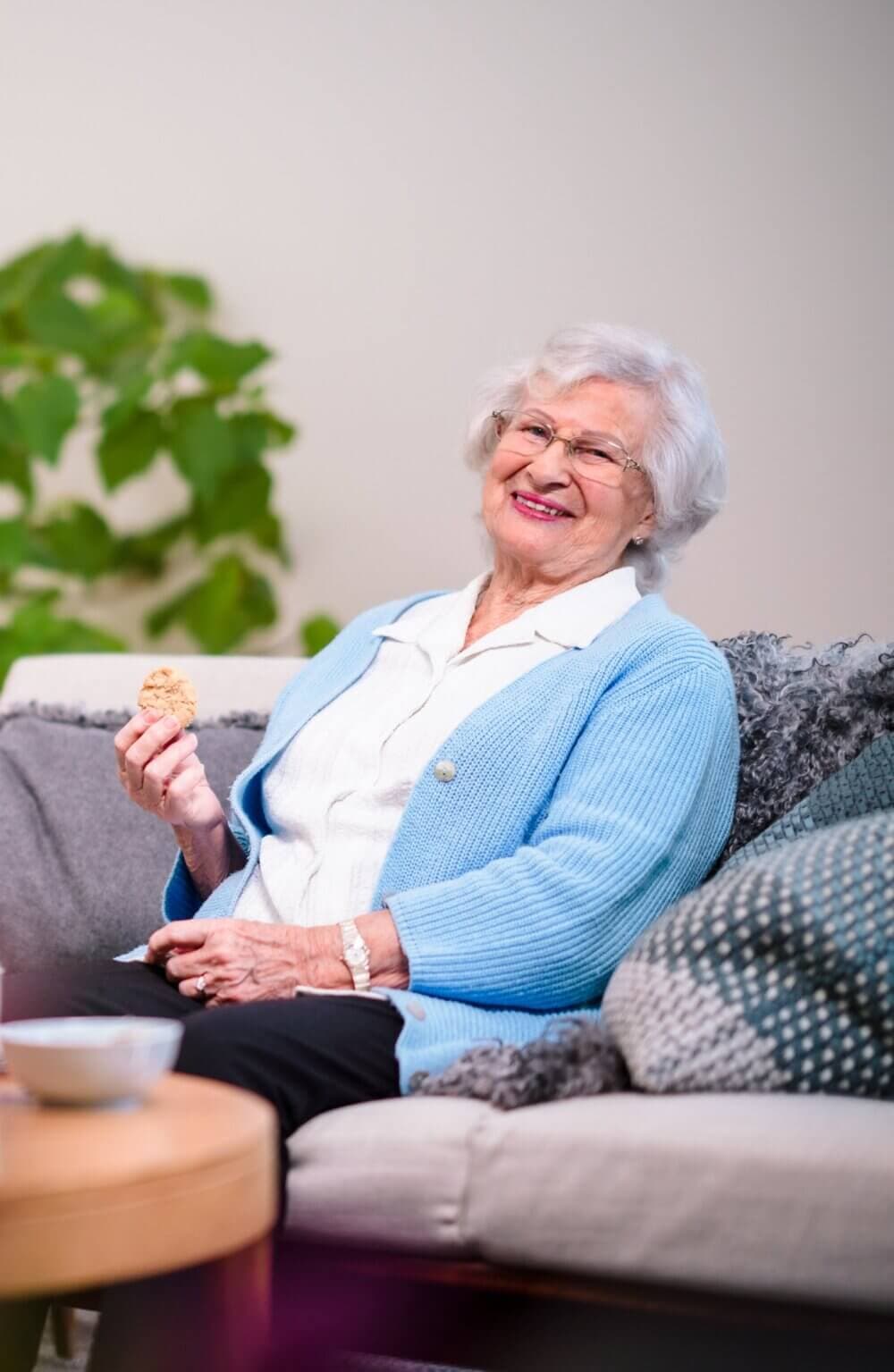 Elderly woman with white hair, blue cardigan, and glasses smiling while holding a cookie and sitting on a couch. - Home Instead