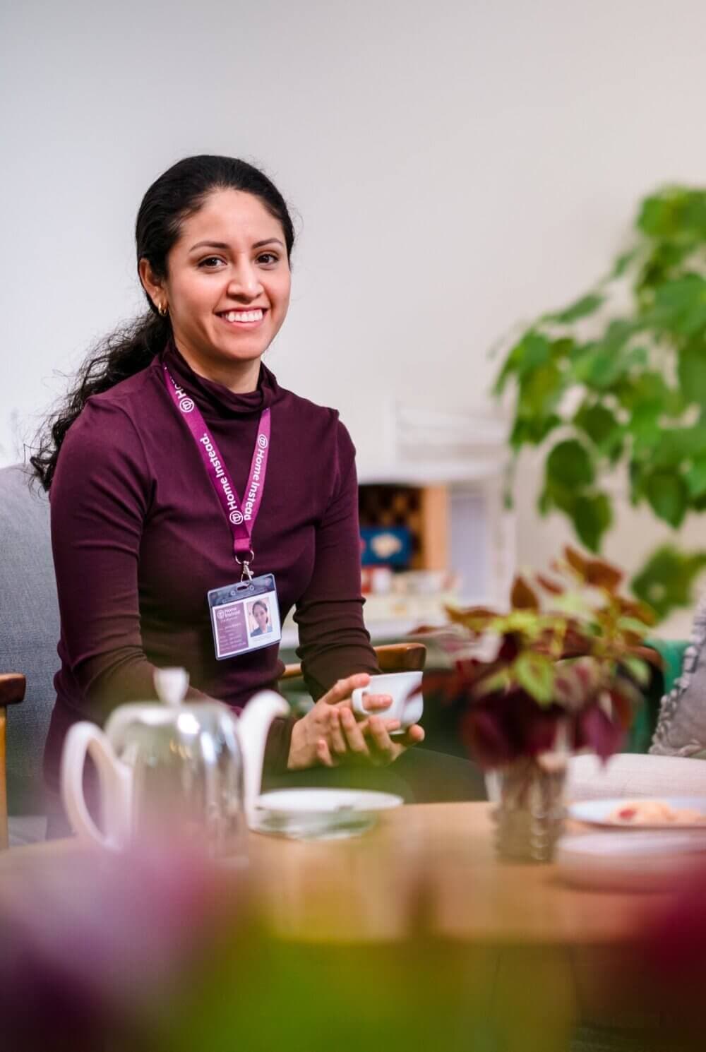 A woman in a maroon sweater, wearing an ID badge, smiles while holding a cup. Tea set on a table and plants in the background. - Home Instead