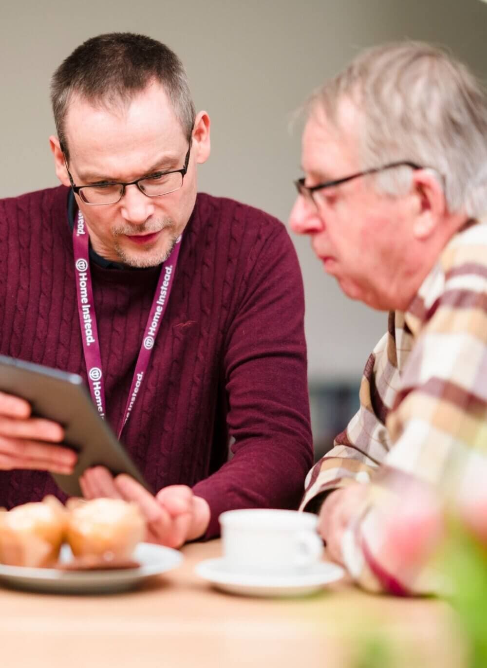 Two men sitting at a table, one showing the other something on a tablet. Coffee and pastries are on the table. - Home Instead