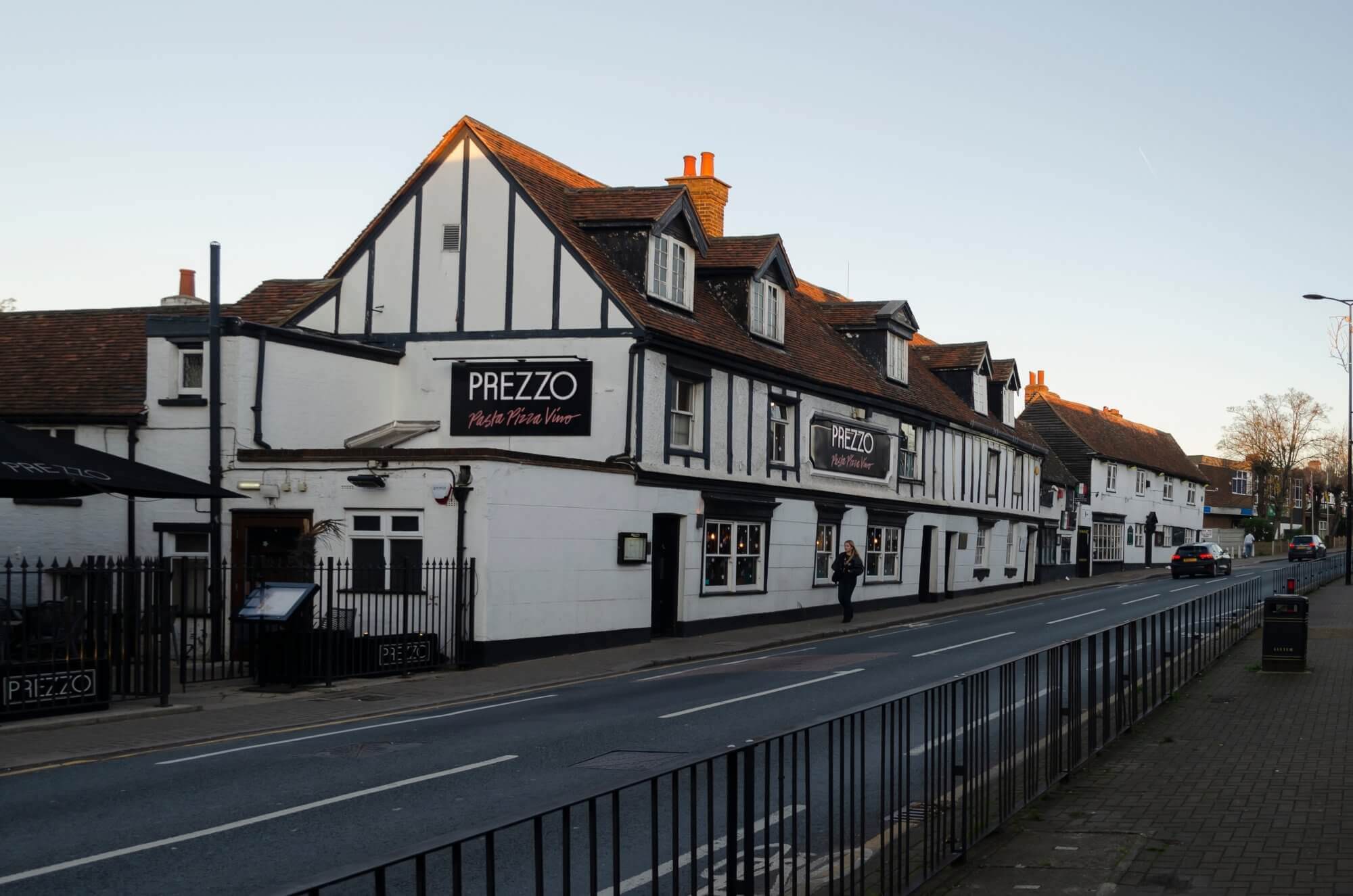 A long building with white walls, black trim, and a sign reading "PREZZO" located along a street with sidewalk fencing. - Home Instead