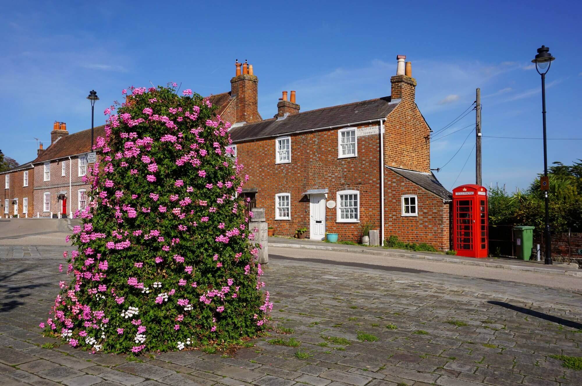 A street with brick houses, a red phone booth, and a large bush of pink flowers in the foreground under a blue sky. - Home Instead Southampton