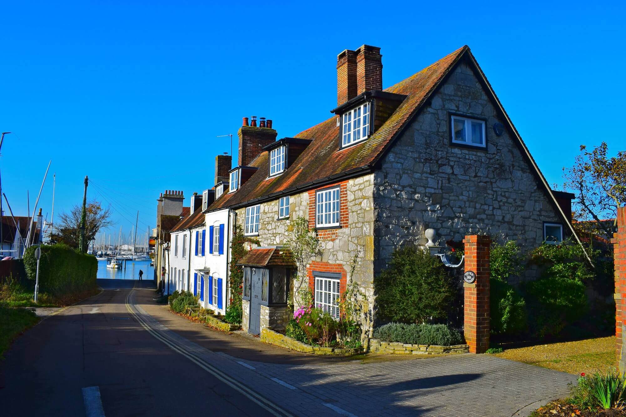 Charming stone cottage along a narrow street, with a marina and sailboats in the background under a clear blue sky. - Home Instead Southampton