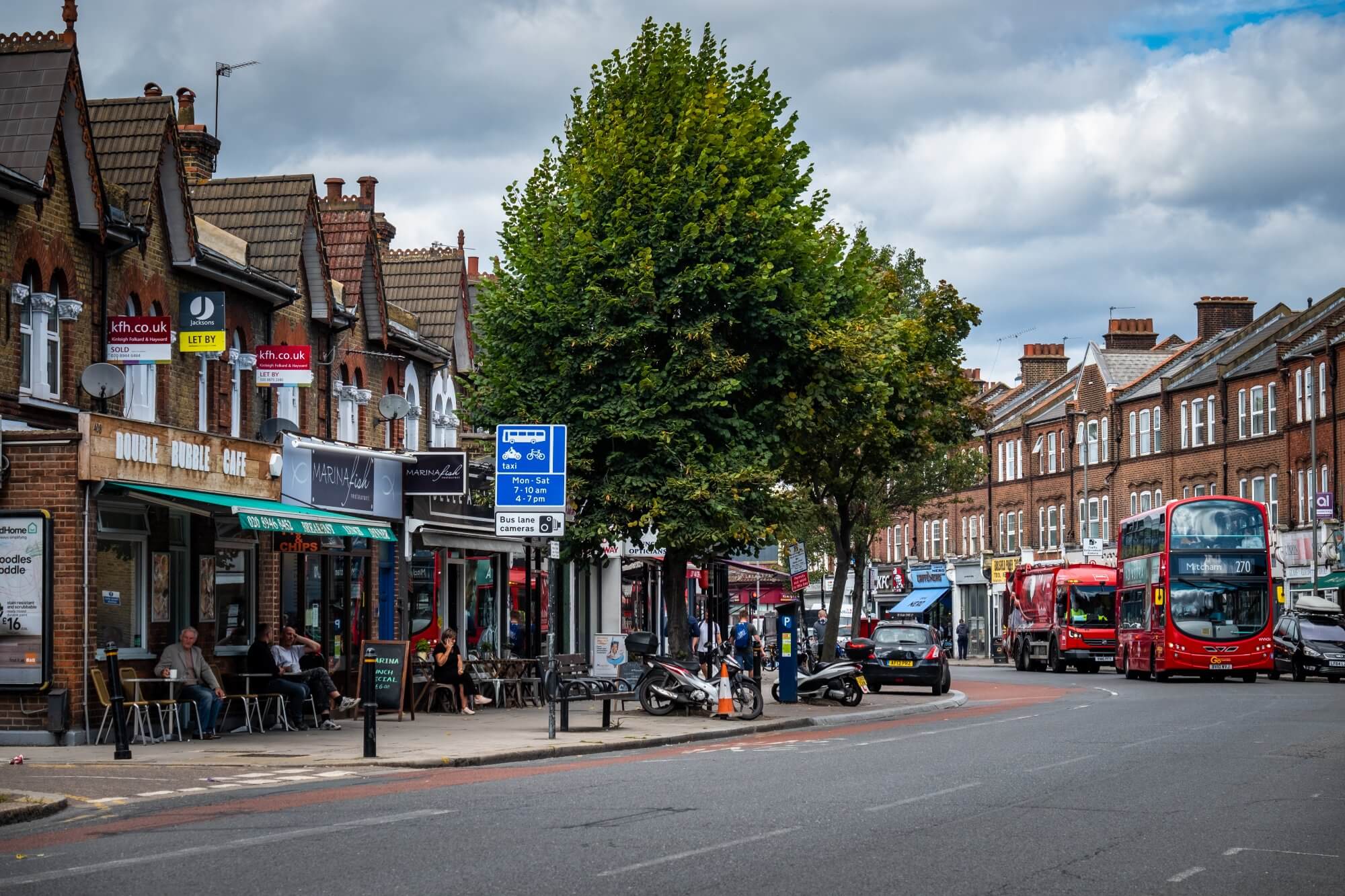 Street scene with shops, cafes, parked cars, and people on the sidewalk. A red double-decker bus is also in view. - Home Instead