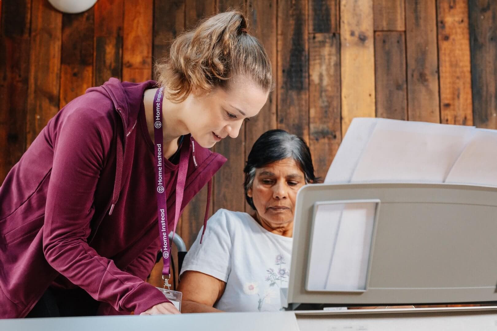 A young woman assists an older woman reading a large document in a brightly-lit, wood-paneled room. - Home Instead