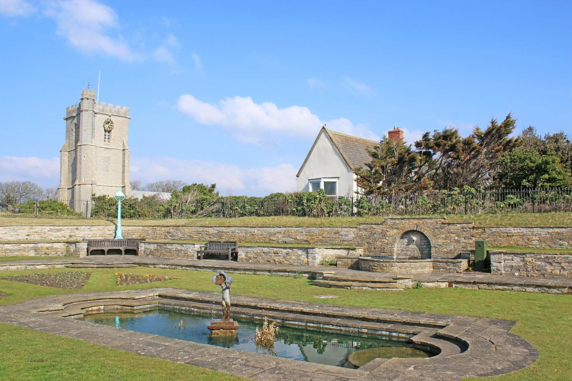 A stone sculpture stands in a rectangular pond, with a church tower and house in the background, set against a blue sky. - Home Instead