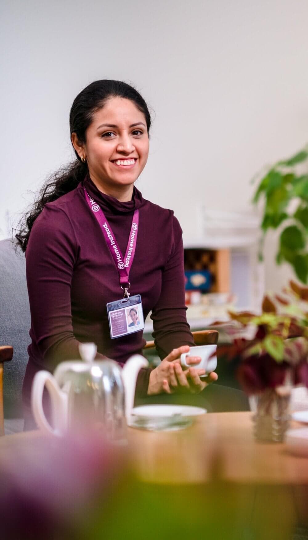 Smiling woman with ID badge holding a cup, seated at a table with a teapot and plants in the foreground. - Home Instead