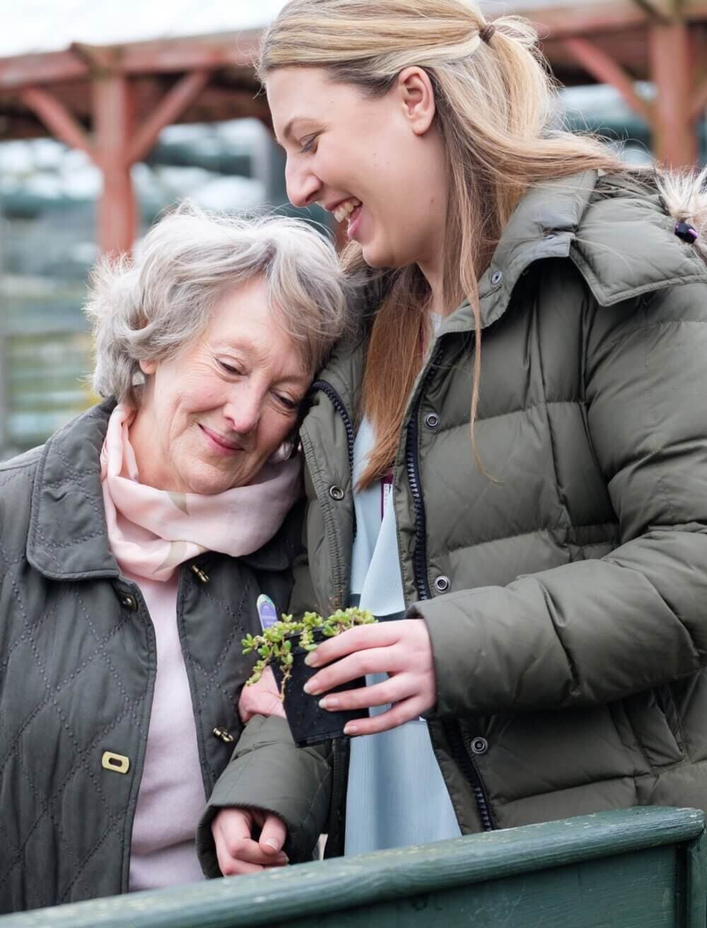 An elderly woman and a younger woman share a joyful moment holding a small plant together. - Home Instead