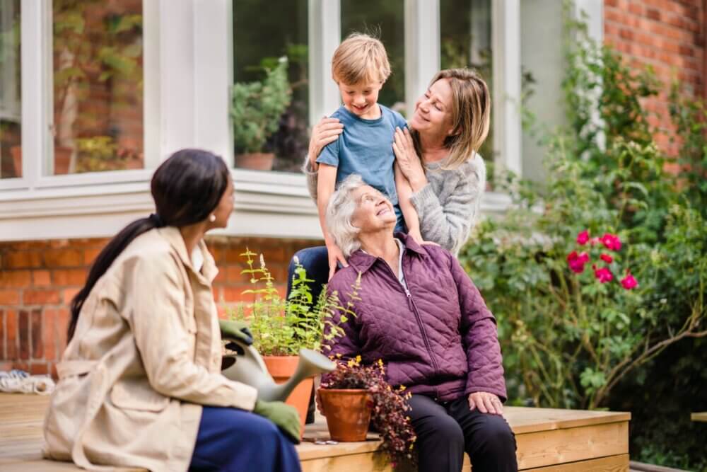 A young boy stands between two seated women with a standing woman beside them; all smiling in a garden. - Home Instead