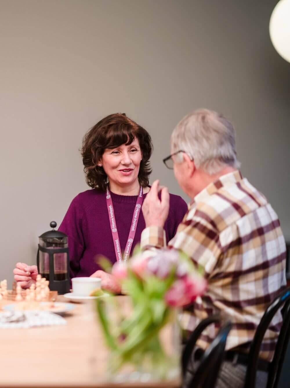 A woman and an older man enjoy a conversation at a table with a French press and flowers. Chess pieces are visible on the table. - Home Instead