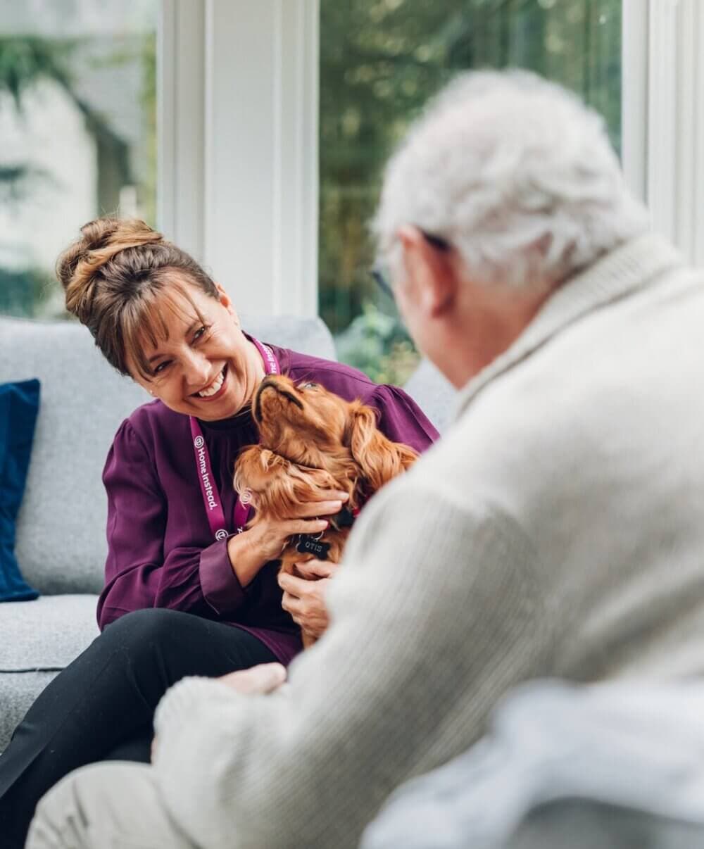 A woman smiles while holding a dog, sitting on a couch beside an elderly man in a white sweater. - Home Instead Southampton