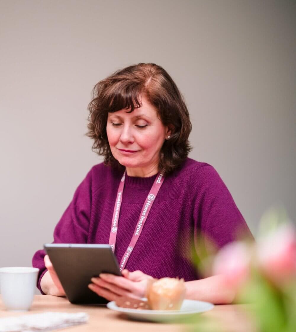 A woman in a purple sweater reads a tablet at a table with a cup, plate, and blurred flowers in the foreground. - Home Instead