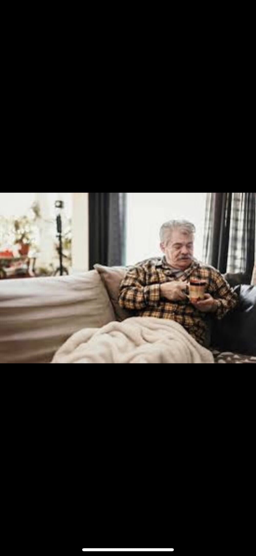 An elderly man with gray hair sits on a sofa, covered in a blanket, holding a mug in both hands. - Home Instead