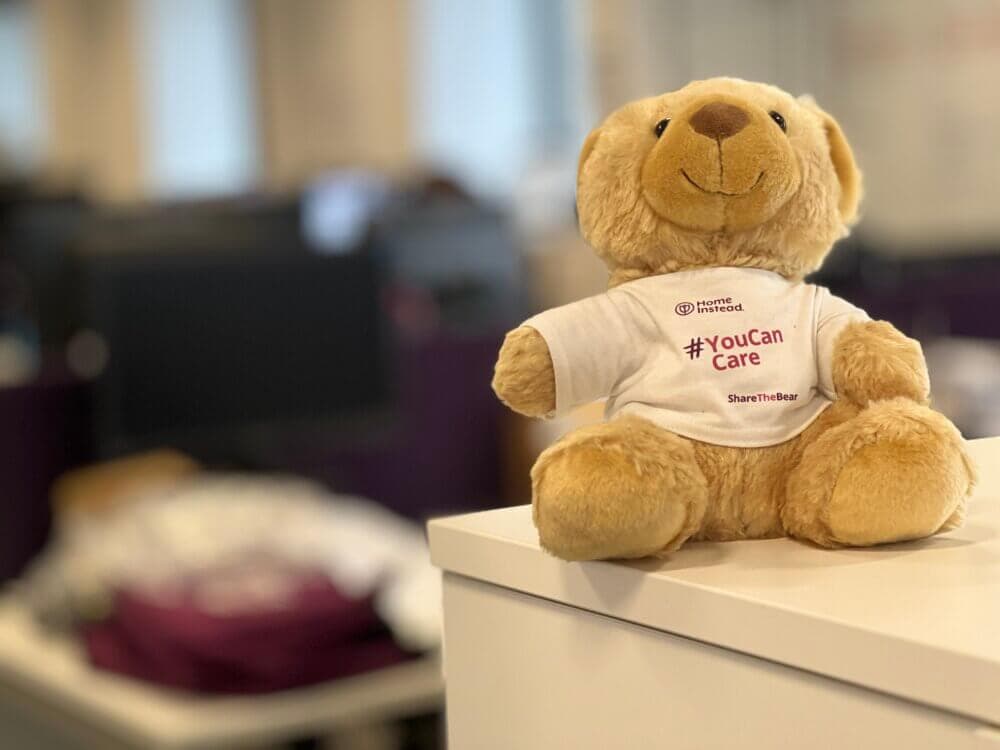 A teddy bear wearing a "#YouCanCare" shirt sits on an office desk with blurred background. - Home Instead