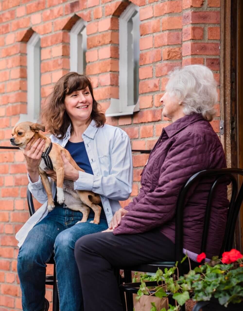 Two women, one holding a small dog, smile while sitting outside a brick building with potted flowers. - Home Instead