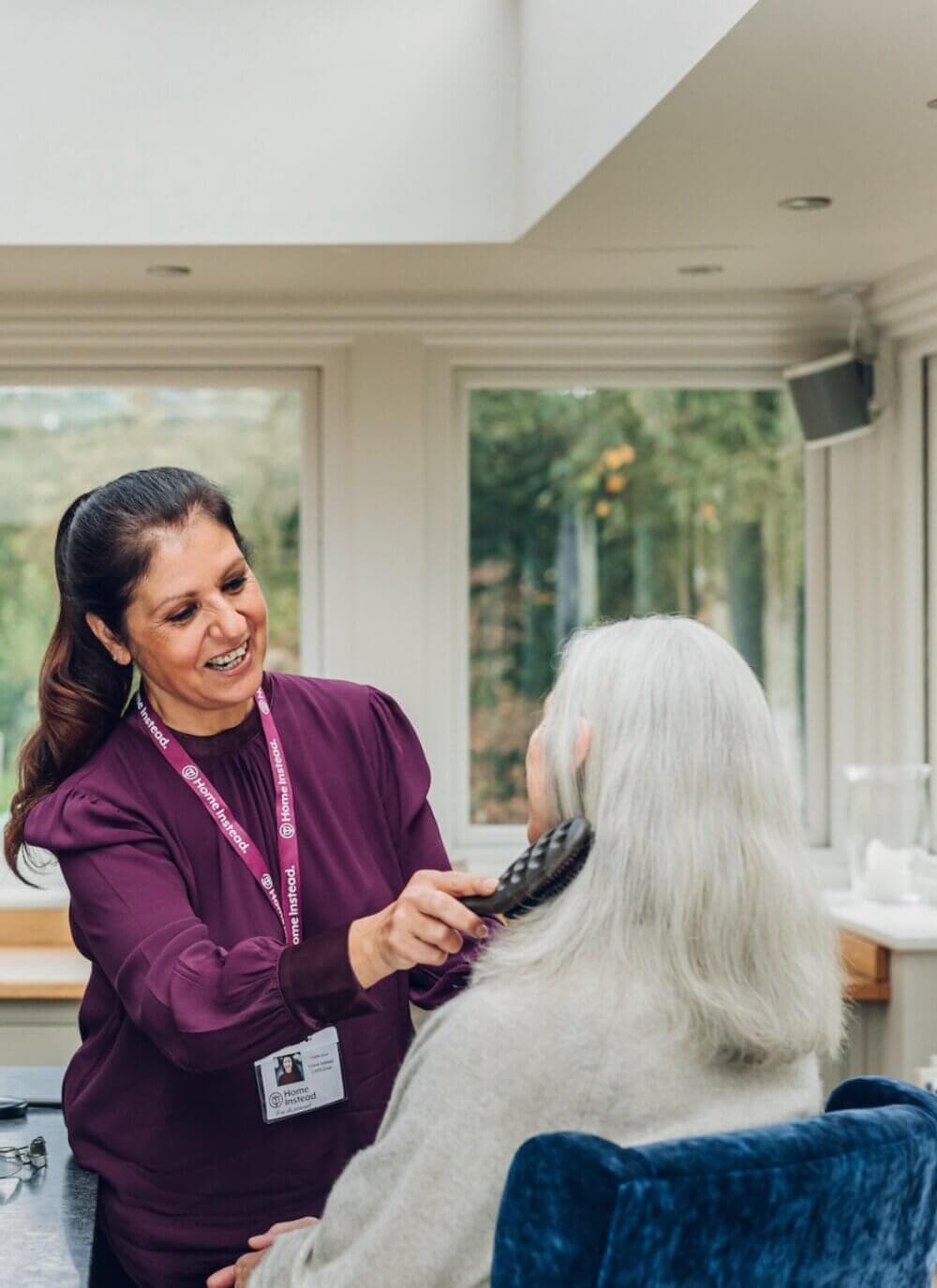 A caregiver in a maroon uniform brushes an elderly woman's white hair in a well-lit room with large windows. - Home Instead