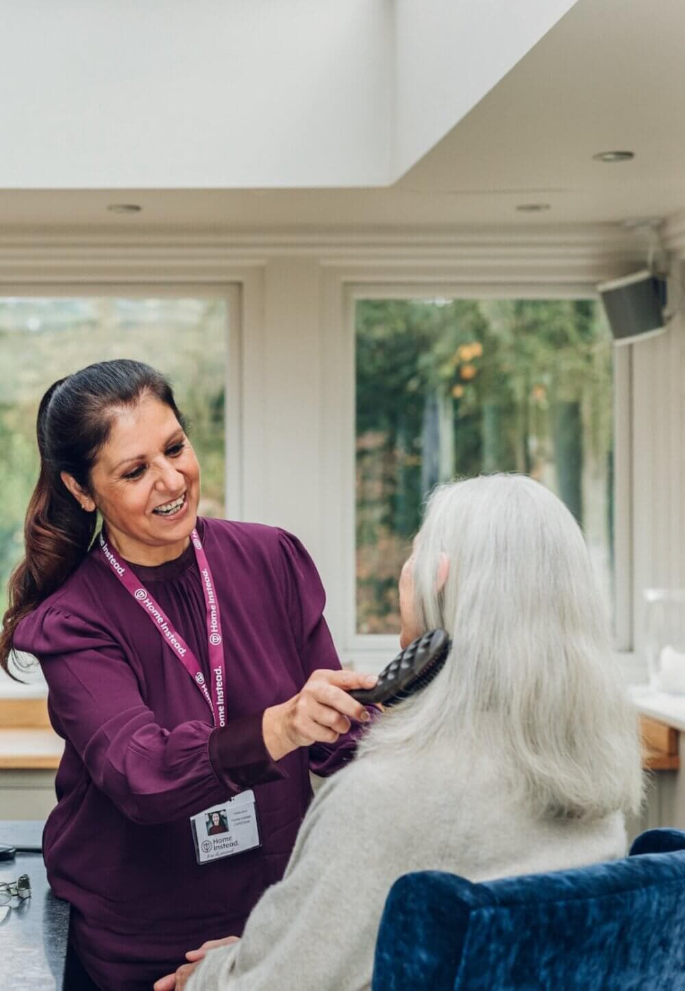 A caregiver smiles while brushing the hair of an older person in a bright room with large windows. - Home Instead