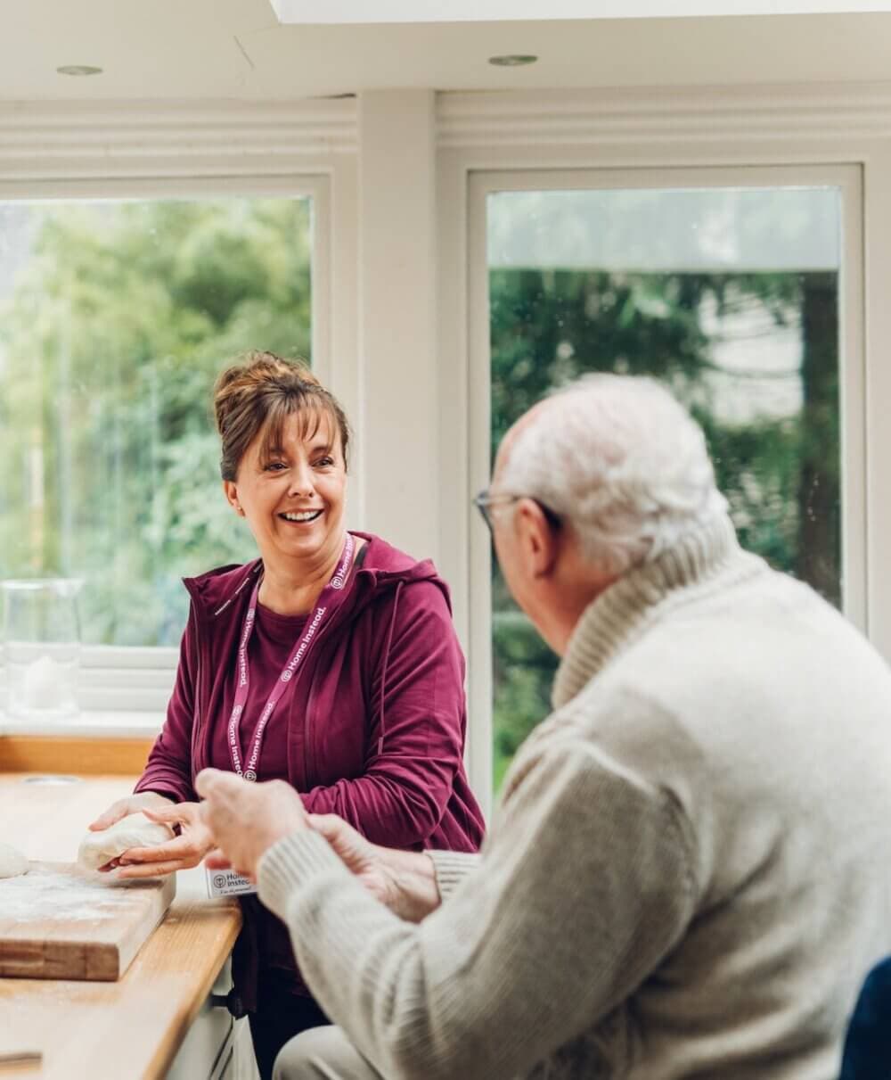 A caregiver and an elderly person sit at a table, smiling and talking, with windows showing greenery in the background. - Home Instead