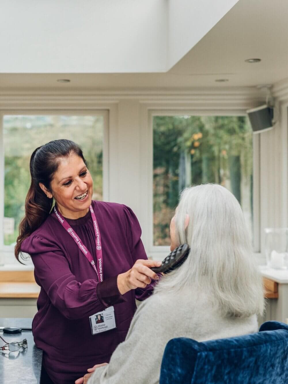 A smiling caregiver brushes an elderly woman's hair in a brightly lit room with large windows. - Home Instead