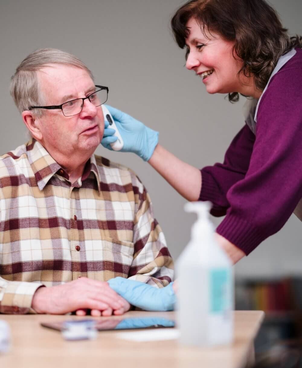 Smiling caregiver in gloves gently cleaning an elderly man's face with a wipe at a table. - Home Instead