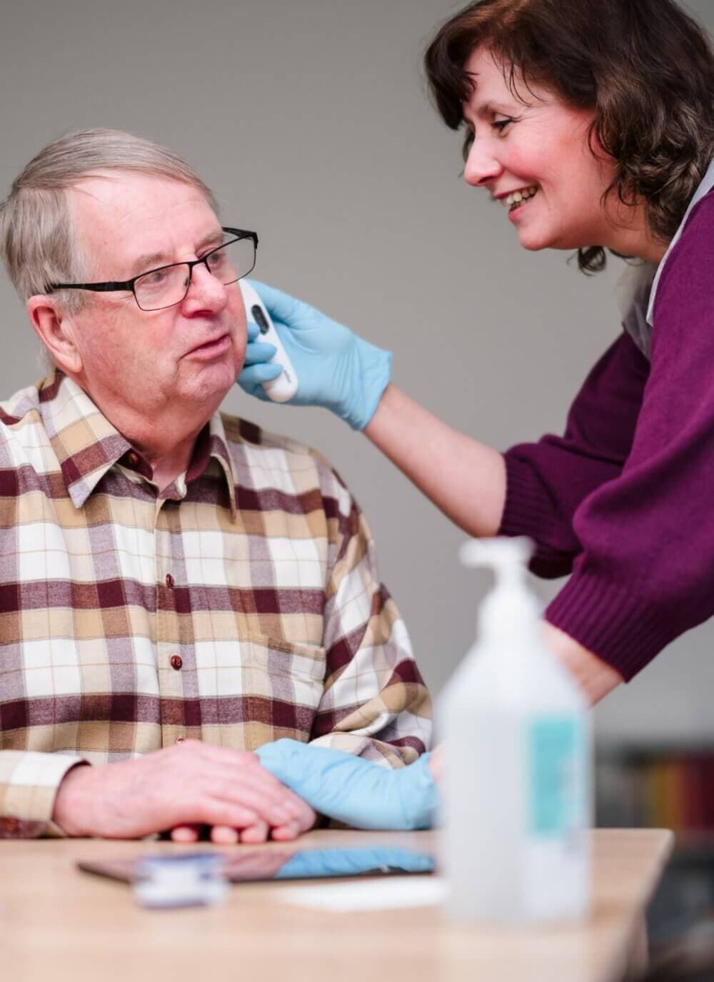 A caregiver wearing gloves checks the ear of an elderly man using a medical device. - Home Instead