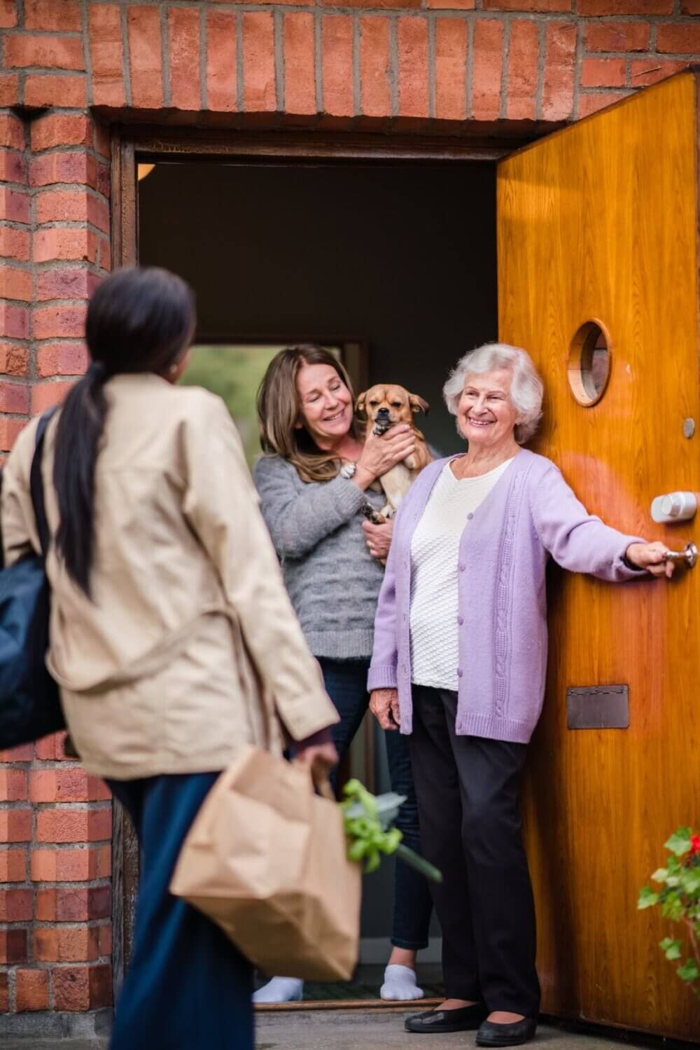 Two women greet a visitor at the door; one holds a dog while the other smiles and opens the door. - Home Instead