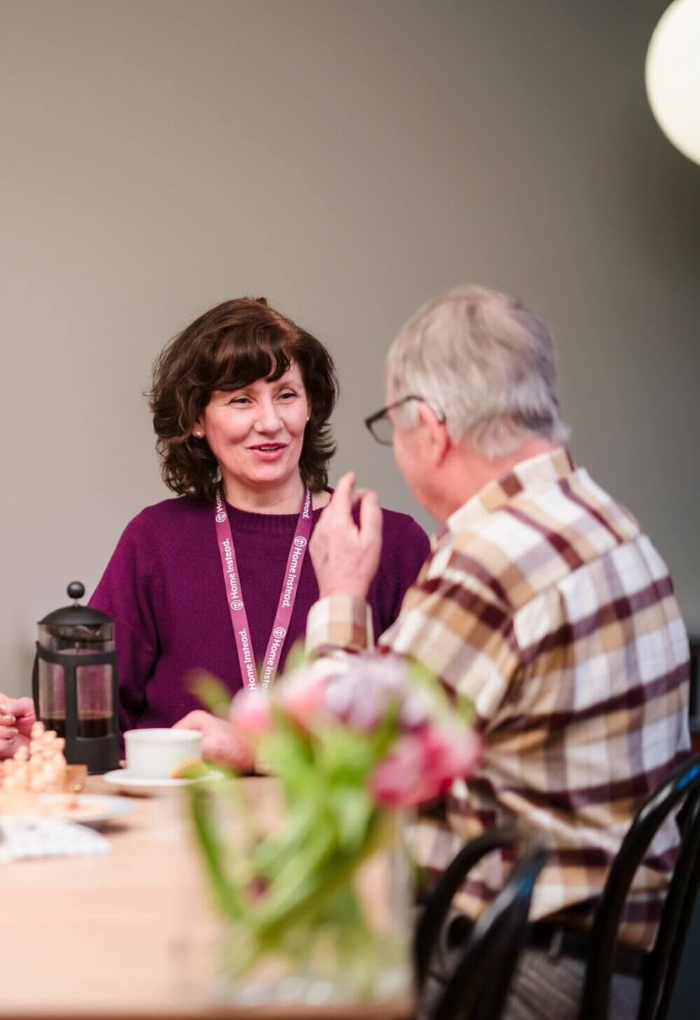 Two people sitting at a table, engaged in conversation, with a French press and flowers in the foreground. - Home Instead