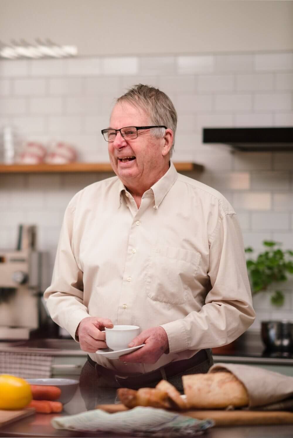 Smiling elderly man with glasses holding a cup in a kitchen, with a table of bread and vegetables in the foreground. - Home Instead