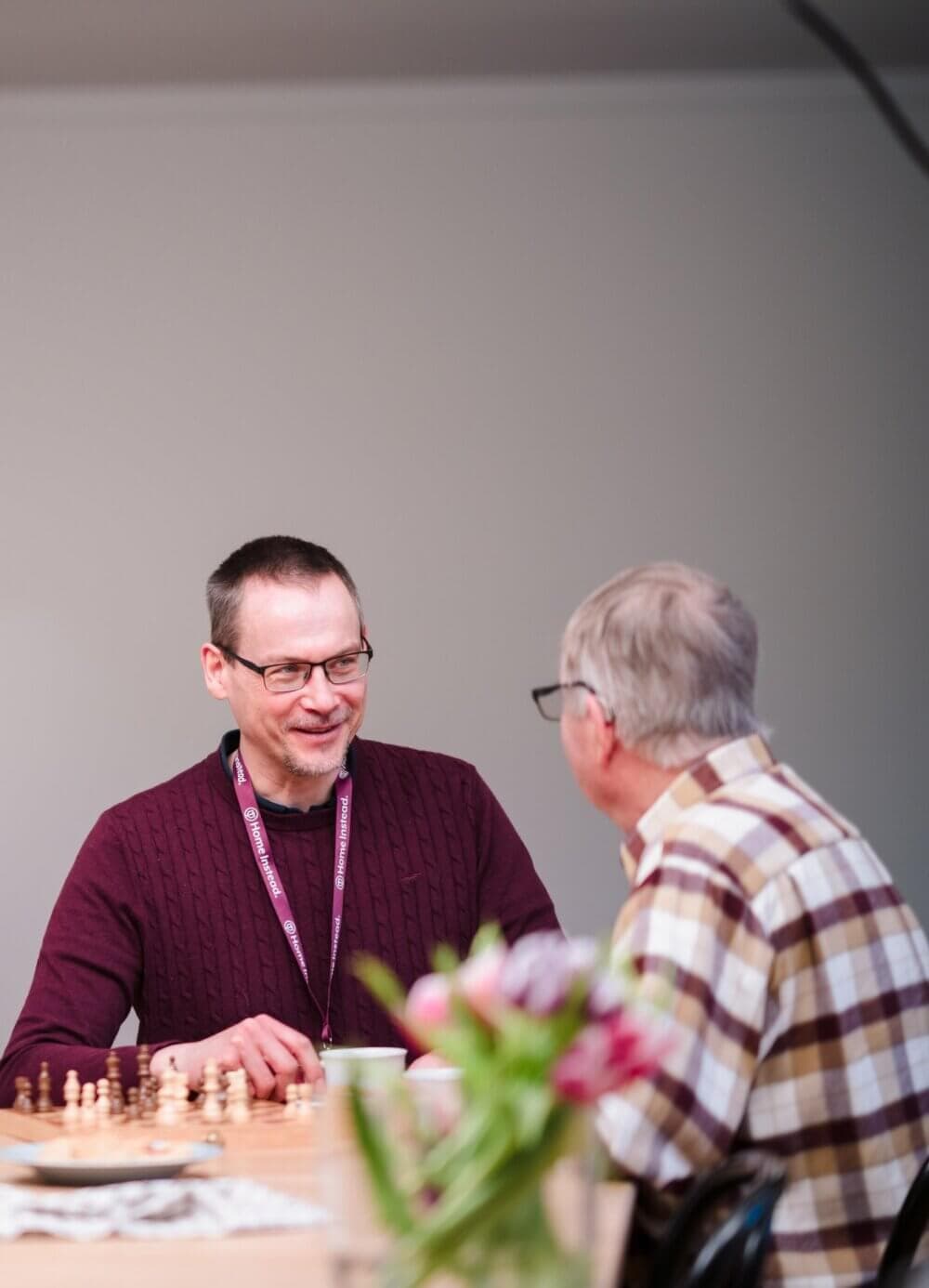 Two men sitting at a table playing chess, engaged in conversation, with a blurred vase of flowers in the foreground. - Home Instead