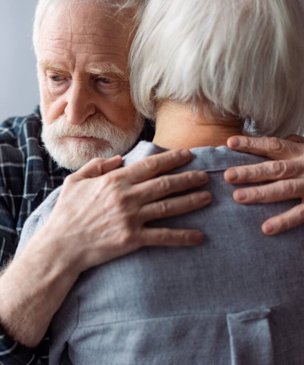 Elderly man with white hair and beard hugging an elderly woman, displaying a tender and comforting embrace. - Home Instead