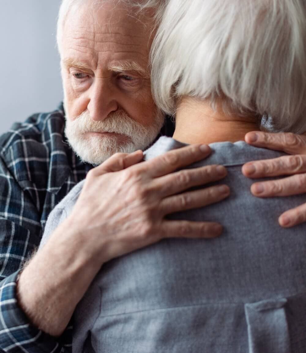 Elderly man embracing an elderly woman, both with white hair, showing support and comfort. - Home Instead
