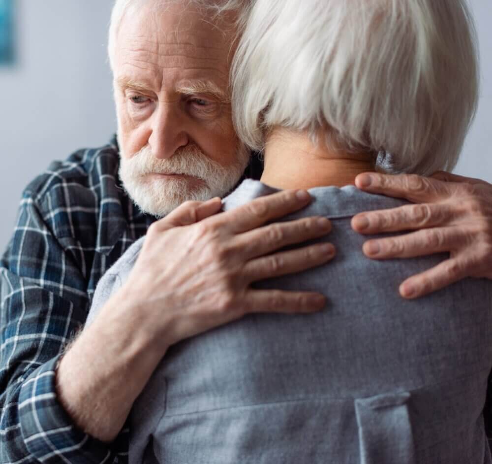 Elderly man with a sad expression hugs an elderly woman, showing emotional support. Their heads are close together. - Home Instead