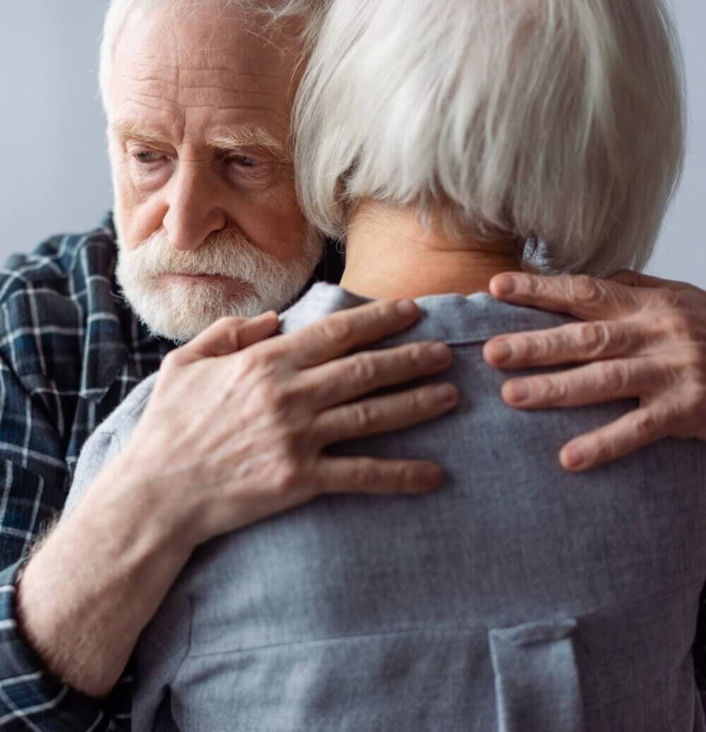 An elderly man with a white beard embraces an elderly woman with short white hair, both wearing gray clothing. - Home Instead