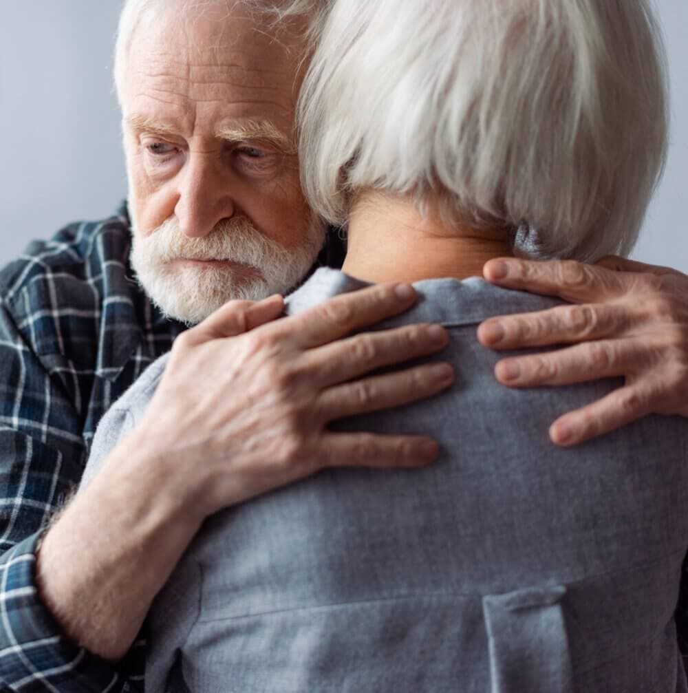 An elderly man with white hair hugs an elderly woman with short white hair, both showing a sense of comfort and support. - Home Instead