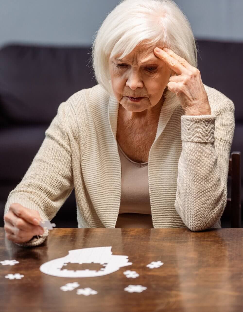 Elderly woman with white hair solving a jigsaw puzzle while sitting at a wooden table. - Home Instead