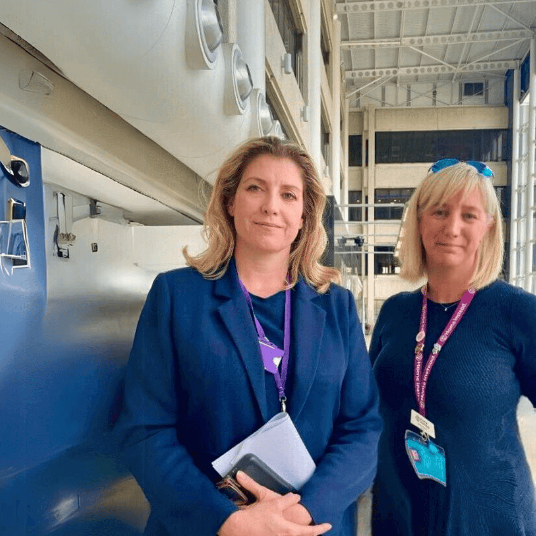 Two women stand in a modern building, each wearing lanyards, one in a blue blazer and the other in a blue sweater. - Home Instead